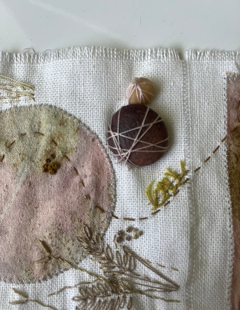 Textile art piece with embroidered elements and natural materials on a neutral background